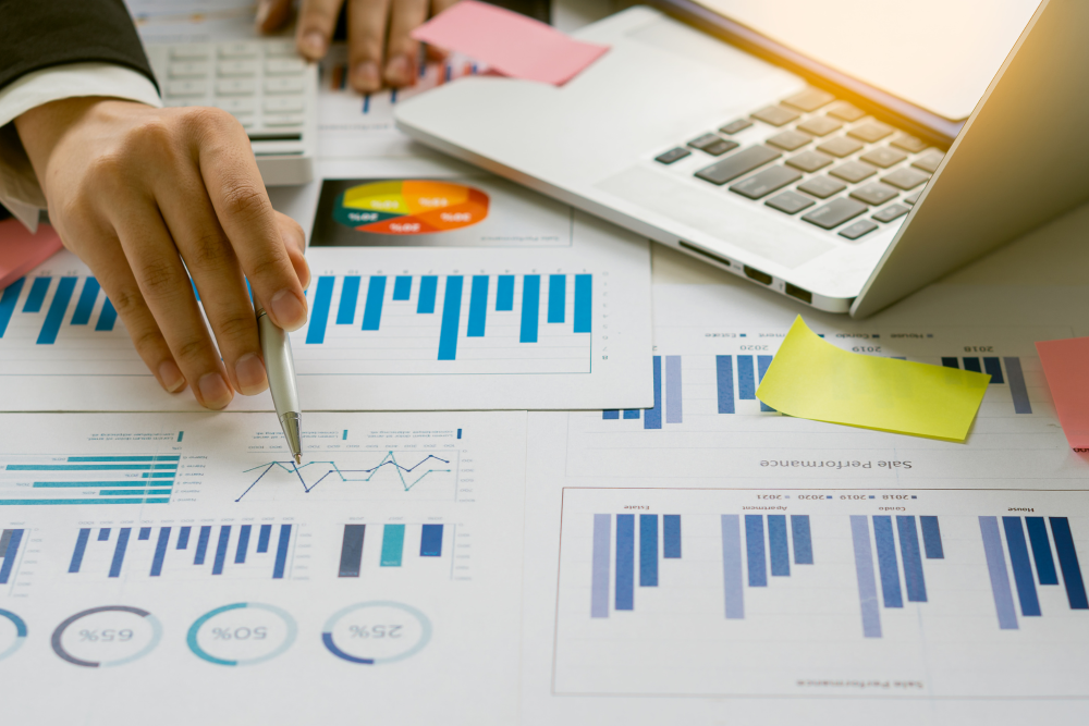 A businessperson is at a desk, pointing at a chart with a pen. Their computer is open and they are working on business reports.