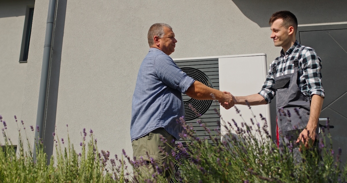 A technician and a homeowner exchange a handshake next to an external heat pump unit in a suburban setting, marking the completion of a maintenance or installation service.