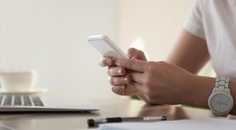 Close up image of modern cellphone in womans hand at desk with laptop. Office worker boring at workplace and playing games, writing in social networks. Businesswoman looking important contact on phone