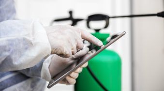 Man in virus protective suit and mask looking and typing on tablet, disinfecting buildings of coronavirus with the sprayer. Infection prevention and control of epidemic. World pandemic.