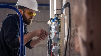 A male electrician works in a switchboard with an electrical connecting cable, connects the equipment with tools.