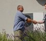 A technician and a homeowner exchange a handshake next to an external heat pump unit in a suburban setting, marking the completion of a maintenance or installation service.