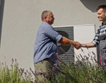 A technician and a homeowner exchange a handshake next to an external heat pump unit in a suburban setting, marking the completion of a maintenance or installation service.
