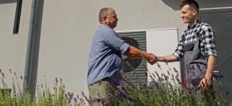 A technician and a homeowner exchange a handshake next to an external heat pump unit in a suburban setting, marking the completion of a maintenance or installation service.