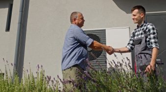 A technician and a homeowner exchange a handshake next to an external heat pump unit in a suburban setting, marking the completion of a maintenance or installation service.