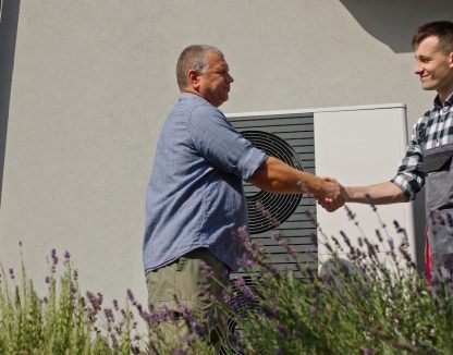 A technician and a homeowner exchange a handshake next to an external heat pump unit in a suburban setting, marking the completion of a maintenance or installation service.